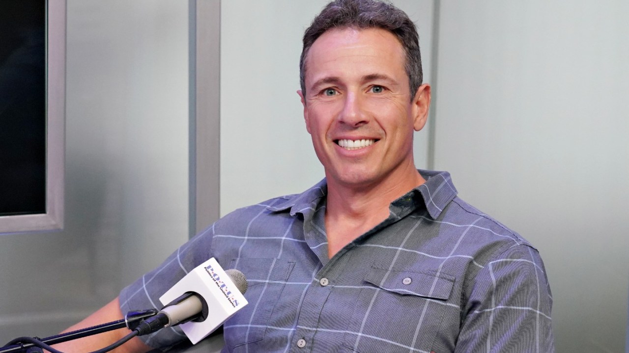 chris cuomo sits in front of a mic, smiling, with a short sleeve button down shirt