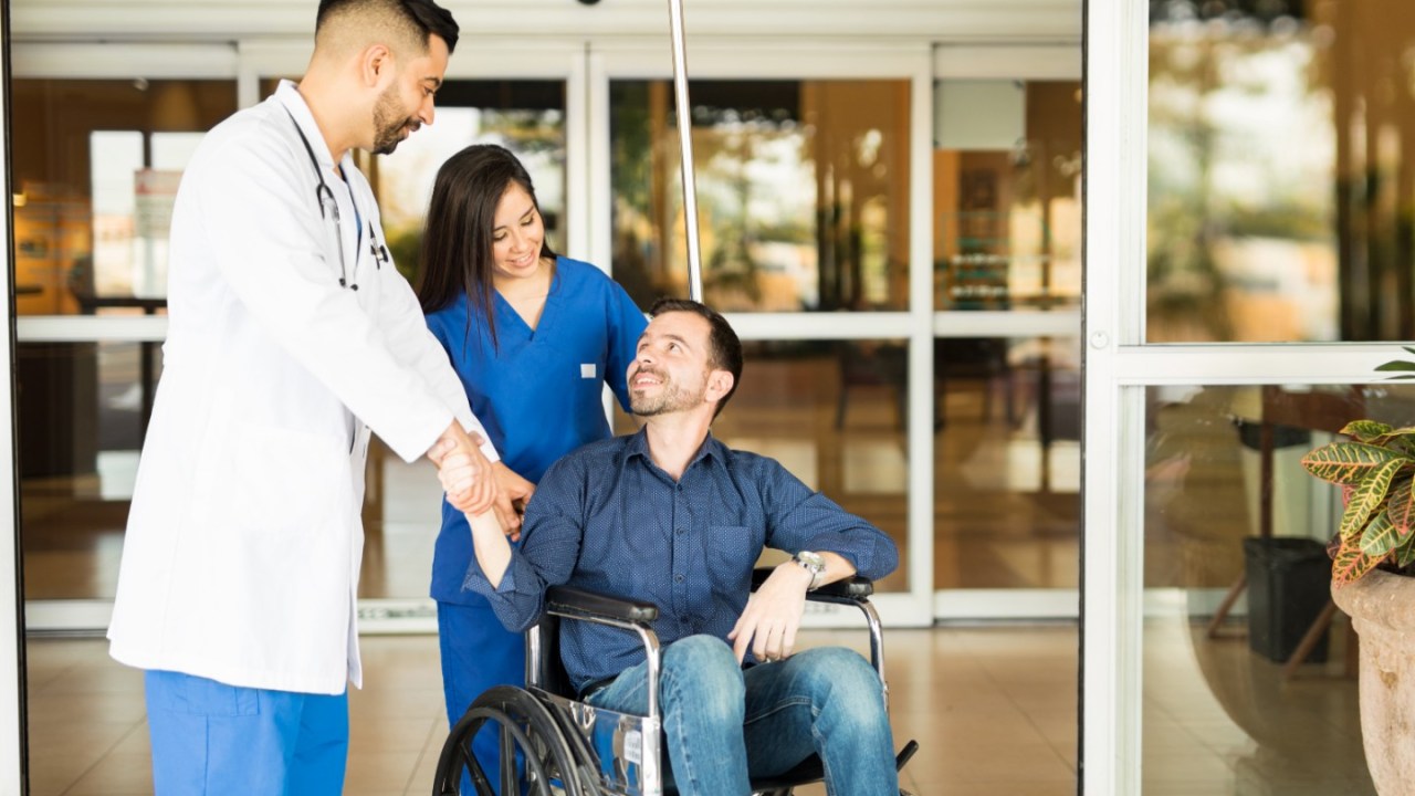 a young female nurse pushes a young male patient out of the hospital on a wheelchair, stopping to speak to a doctor