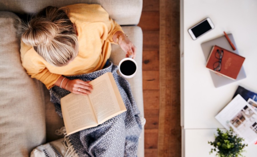 a blonde woman sits on the couch with a drink in a cup and a book and her phone and laptop closed on the coffee table