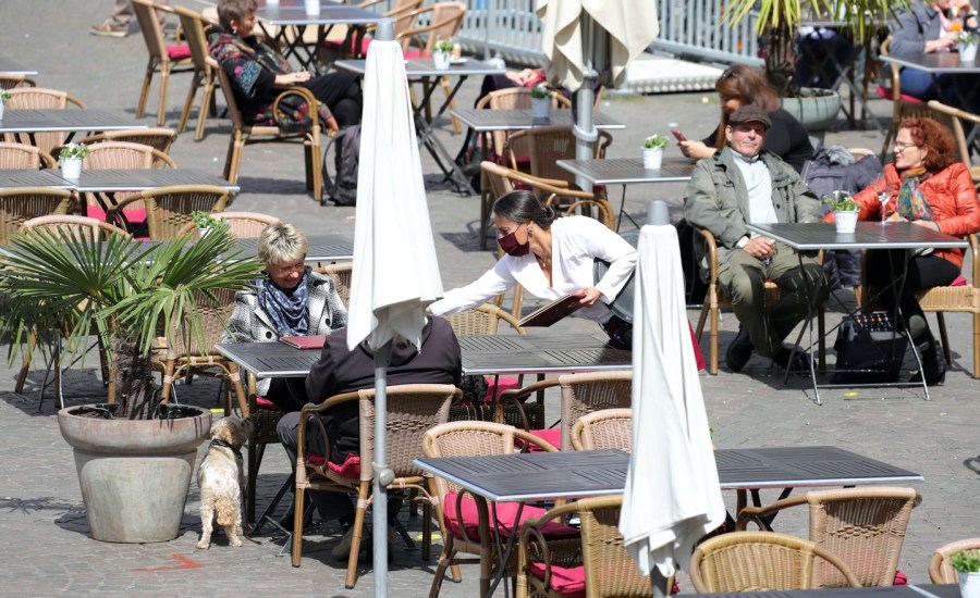 people wear masks and sit at outdoor tables of a restaurant while being served by a waitress