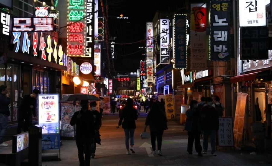 people wearing masks roam around shops and other businesses on a street in South Korea at night