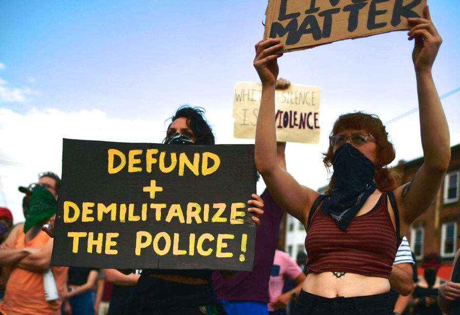 a protestor holds a sign that reads "demilitarize and defund the police" next to another holding a sign that says "black lives matter"