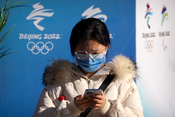 A woman wearing a face mask to protect against COVID-19 sits near landscaping decorated with the logos for the Beijing Winter Olympics and Paralympics