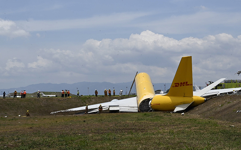 A yellow DHL cargo jet that spun off lays broken on the runway as workers are seen in the distance