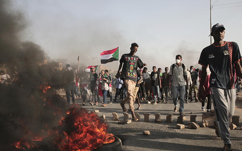 Protestors waving Sudanese flags walk past car tires on fire