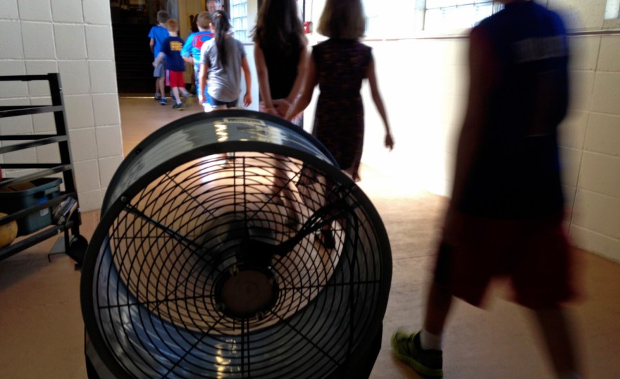 Students walk by fan in school hallway.