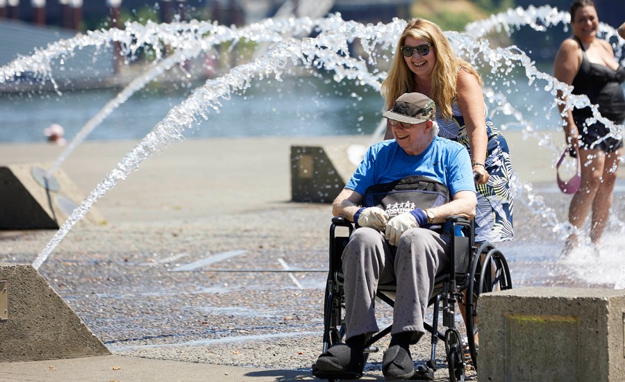 Women pushing man in wheelchair through fountain.
