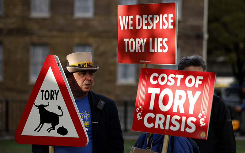 Demonstrators protest opposite Parliament with red and white signs depicting "Tory" and a bull defecating, "We despise Tory lies" and "Cost of Tory crisis"