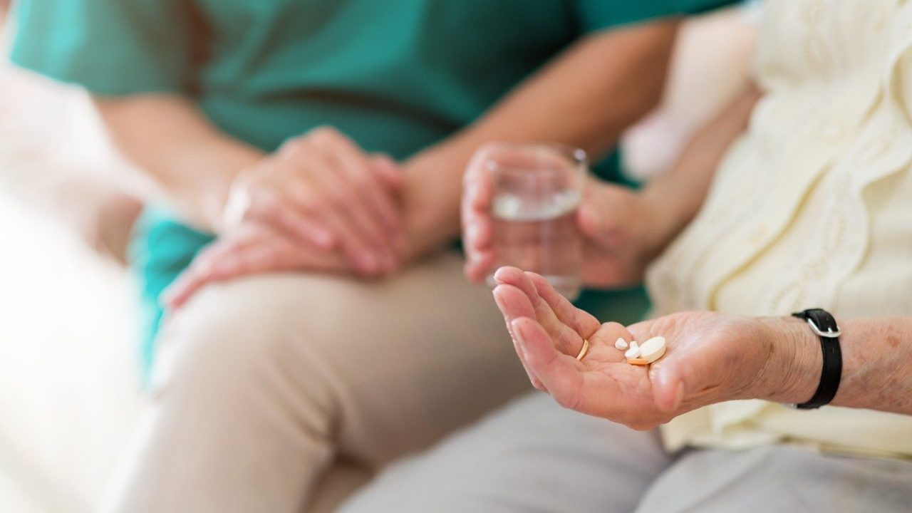 older person with pills in the palm of their hand, seated next to a nurse