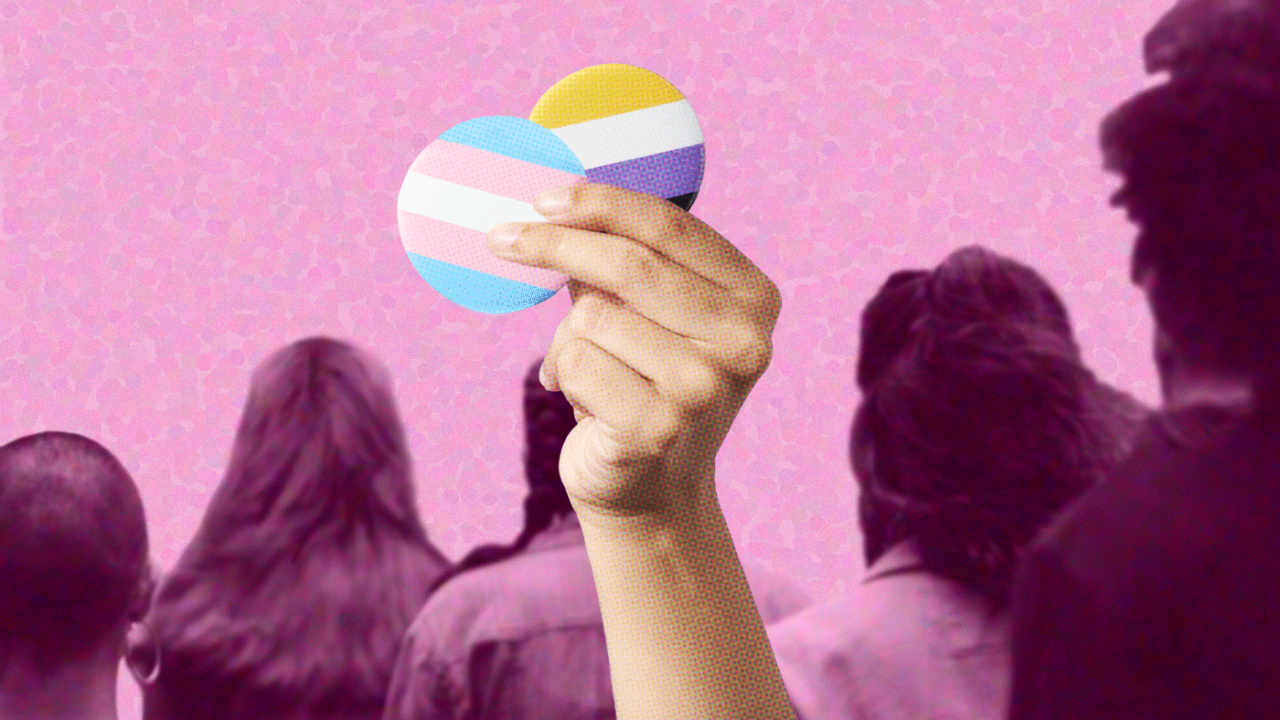 Photo illustration of a person's hand holding two pins, one with transgender flag colors (light blue, light pink, white) and one with non-binary flag colors (yellow, white, purple and black). Hand is over a pink-dotted background with a purple-toned group of people, as seen from behind.