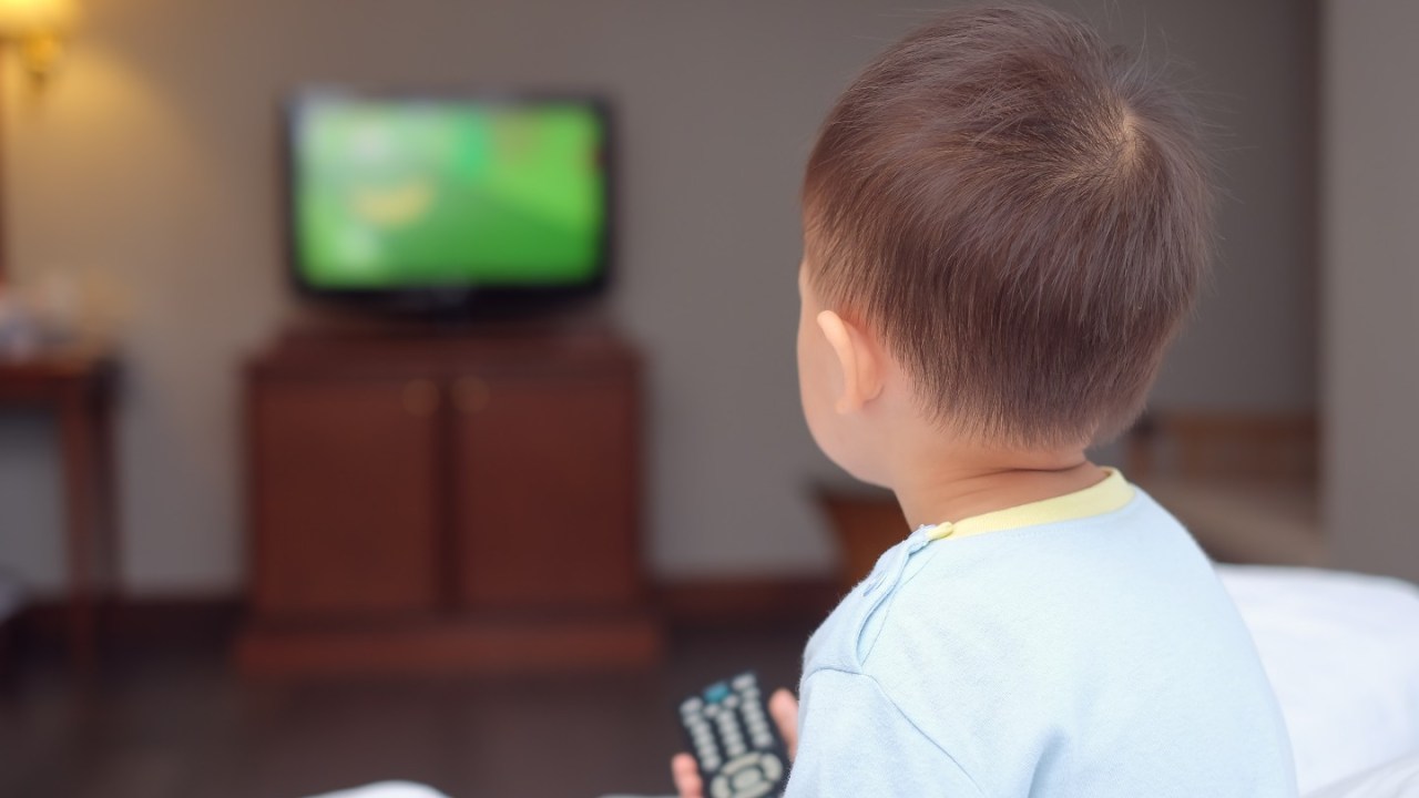 young child sitting on bed facing away at a TV holding a remote controller