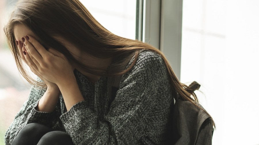 female teen sitting at a window with head in hands, wearing backpack