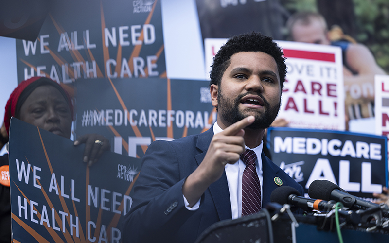 Rep. Maxwell Frost (D-Fla) discuss Medicare for All at a podium as supporters hold signs in the background