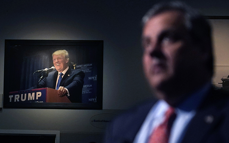 A photograph of former President Trump hangs on the wall in the background while an out-of-focus Chris Christie speaks in the right foreground