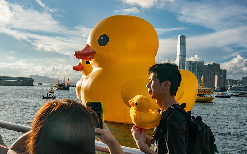 People holding rubber duck stuffed toys pose for photos near giant inflatable rubber duck sculptures in Victoria Harbor