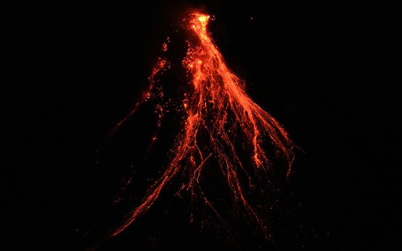 Lava flows down the slopes of Mayon Volcano; the background and volcano are very dark, showing only the orange and red glow of flowing lava.