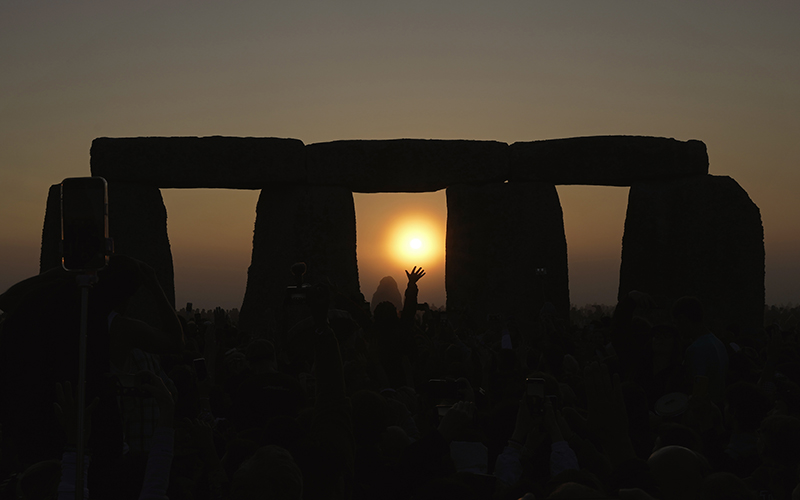 The sun sets at Stonehenge during the summer solstice. The sky is dimming and the sun is framed in one of the openings in Stonehenge, while the silhouette of a reveler's hand is seen reaching up toward the sun.