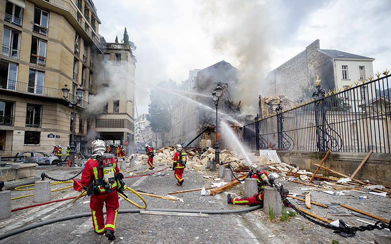 Firefighters try to extinguish a Thursday fire at the site of an explosion in Paris. Rubble fills the street and smoking buildings are on the left and right of the street. Firefighters are seen carrying hoses and spraying water at the buildings