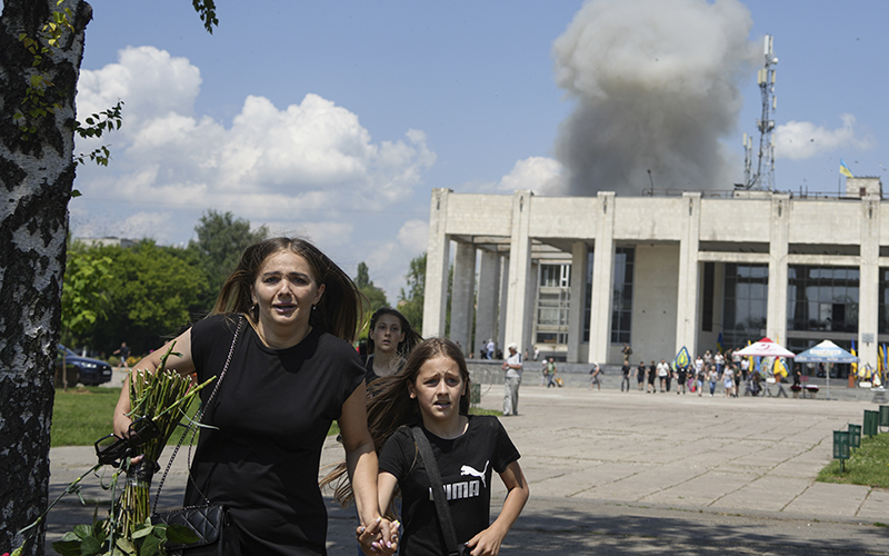 A family runs from an explosion after a Russian rocket attack. The woman is clutching her child's hand as the run toward the viewer, while behind them is seen a white building with smoke rising above it.