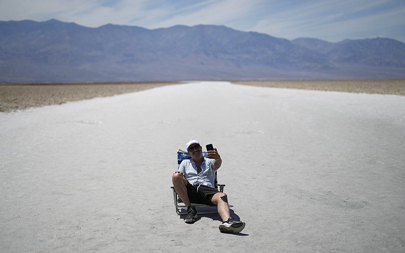 A man sits in a low chair as he takes a selfie on the salt flats at Badwater Basin in Death Valley National Park. The white ground that his chair is on is seen extending away into the distance, where is also see a faraway mountain range.