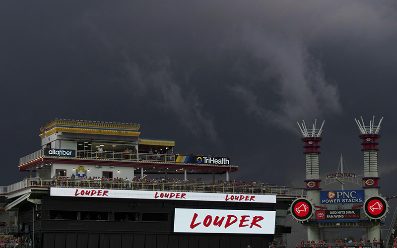 Storm clouds are seen behind Great American Ballpark during a baseball game. The sky is dark and ominous behind a section of the park where there is a railing with fans lined up. Below where the fans stand are digital signs that read "LOUDER"