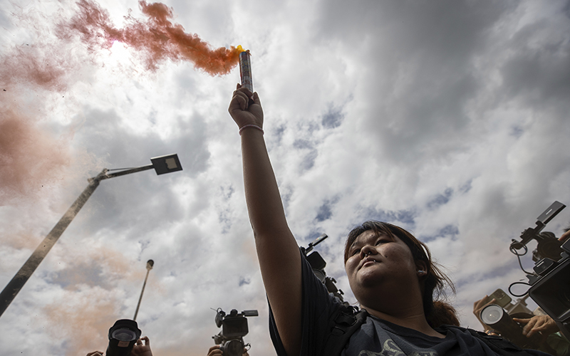 A supporter of Move Forward Party holds a smoke flare during a protest outside Parliament in Bangkok. The angle of the viewer is from below the protester looking up at the sky. The flare is releasing an orange colored smoke, and beyond it is seen an overcast and cloudy sky.