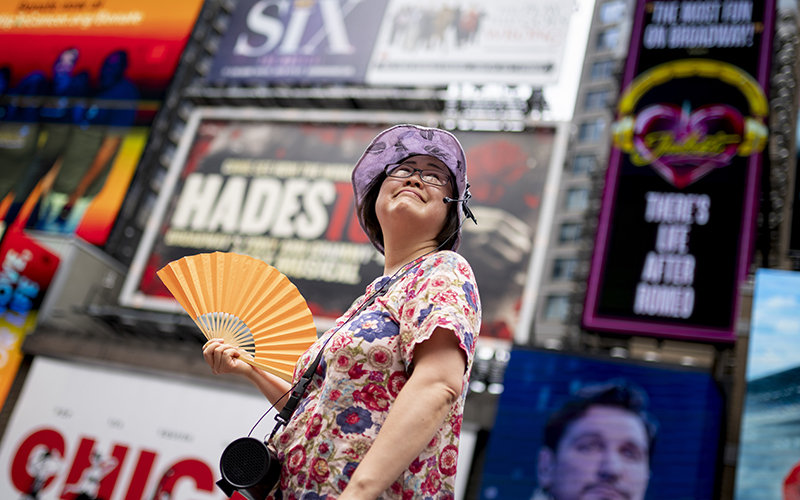 A woman wearing a wide-brimmed hat looks up toward the sky and fans herself as she stands in Times Square.