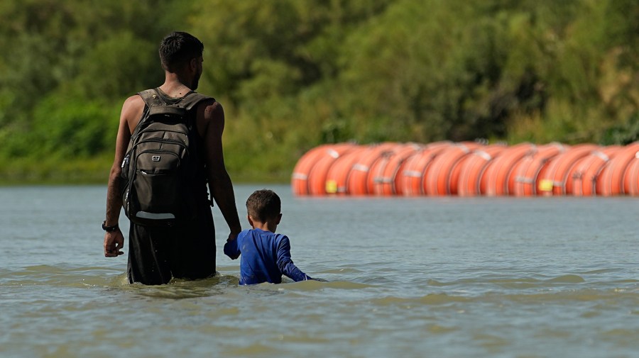 Migrants walk past large buoys being used as a floating border barrier on the Rio Grande Tuesday, Aug. 1, 2023, in Eagle Pass, Texas.