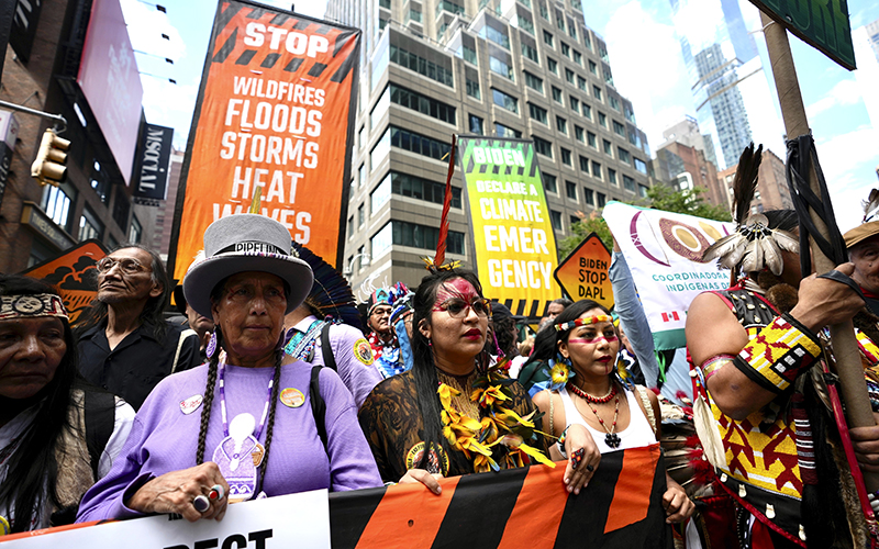 Climate activists march protesting energy policies and the use of fossil fuels. In the front row of protesters are several Indigenous people wearing feathers, beads or face paint. Behind them, people in the crowd carry signs that read "STOP WILDFIRES FLOOD STORMS HEATWAVES," "BIDEN DECLARE A CLIMATE EMERGENCY," and "BIDEN STOP DAPL"