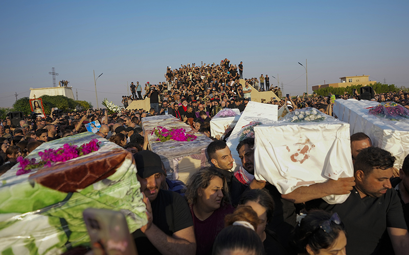 A large crowd of people is gathered in the distance. In the foreground are people carrying coffins draped with fabric and flowers.