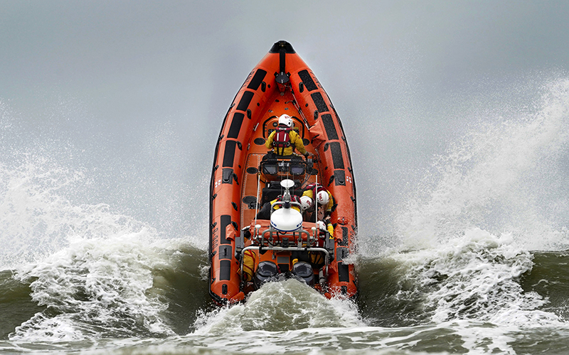 An orange boat crashes through the waves. After hitting a sharp wave, the boat appears to be pointed straight up out of the water, showing the passengers and equipment aboard.