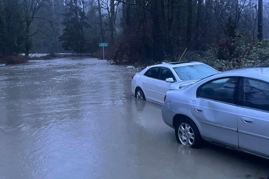 In this photo provided by Kira Mascorella, cars drive through flood waters in Granite Falls, Wash., Tuesday, Dec. 5, 2023. An atmospheric river has brought heavy rain, flooding and warm winter temperatures to the Pacific Northwest, closing rail links, schools and roads as it shattered daily rainfall and temperature records in Washington state. (Kira Mascorella via AP)