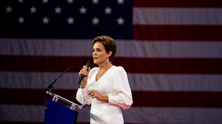 Arizona Republican Senate candidate Kari Lake speaks at a podium against a backdrop of a large American flag.