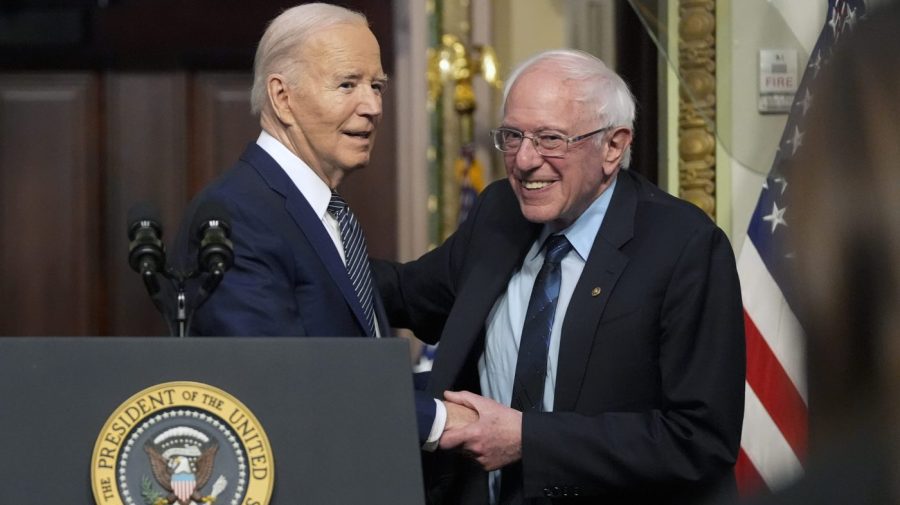 President Biden and Sen. Bernie Sanders embrace and shake hands while giving a speech.