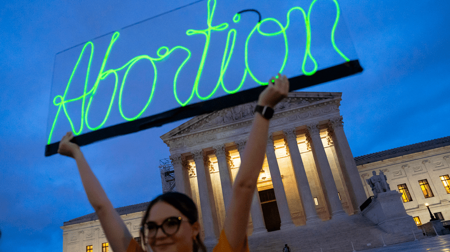 A woman hold up a neon green sign that says abortion in front of the Supreme Court.