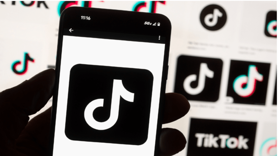 A person holds a phone displaying the TikTok logo against a computer screen showing more logos.