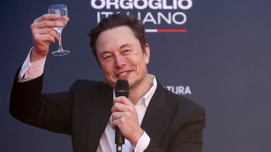 Elon Musk holds up a glass while speaking at an event.