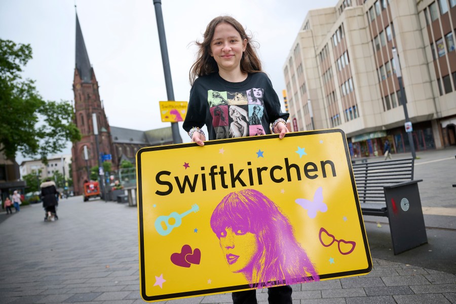 Schoolgirl Aleshanee Westhoff shows a "Swiftkirchen" town sign in honor of musician Taylor Swift in Gelsenkirchen, Germany, Tuesday, July 2, 2024. The Swifties are about to take over the German city of Gelsenkirchen, where American superstar Taylor Swift is set to give three concerts of her Eras Tour later this month. In honor of the singer, the city renamed itself “Swiftkirchen" — at least temporarily to welcome the tens of thousands of fans who are expected to come for the Eras Tour shows on July 17, 18 and 19, German news agency dpa reported. (Bernd Thissen/dpa via AP)