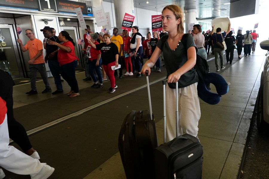 A traveler walks through the departure area as airline catering workers who are employed by Gate Gourmet picket with their supporters, calling for a new union contract with raises and affordable health insurance, Wednesday, July 3, 2024, at Miami International Airport in Miami. A long Fourth of July holiday weekend is expected to create new travel records. (AP Photo/Lynne Sladky)